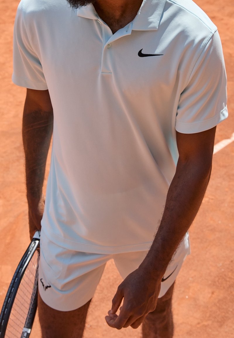 Jasnoniebieska koszulka polo i spodenki z czarnymi akcentami; gładka tkanina; sportowiec trzymający rakietę tenisową na kortie ziemnym.