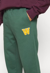 Gröna bomullssweatpants med sidofickor och en broderad gul "W"-logga. Matchas med en vinröd långärmad skjorta.