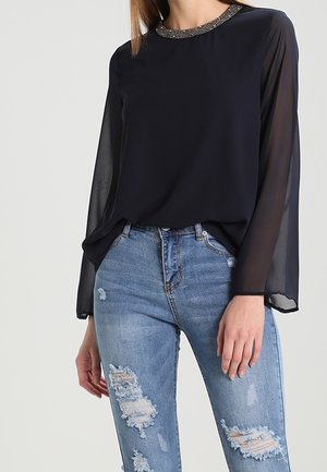Vrouw draagt een zwarte doorschijnende blouse met lange mouwen, versierde halslijn en lichtblauwe gescheurde spijkerbroek.