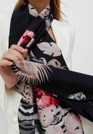 Femme portant un blazer blanc tenant une écharpe noire ornée de grandes feuilles beiges et de motifs floraux rouges près de la poitrine.