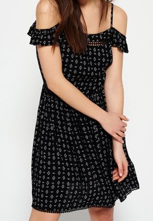 Woman wearing a black off-shoulder dress with white geometric patterns, holding her left arm gently with her right hand.