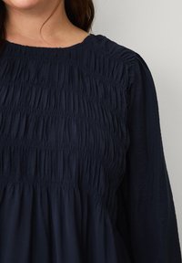 Mørkeblå, smocket langærmet kjole med rund halsudskæring og tekstureret stof, der har fine detaljeindsamlinger og en flydende silhuet.