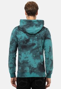 Kapuzensweatshirt im teal und schwarzen Batikmuster. Mit gerippten Bündchen und Saum, einem grafischen Design auf der Rückseite und weichem Stoff.