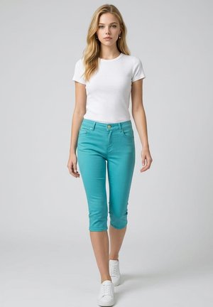 Mujer rubia caminando hacia adelante con una camiseta blanca de manga corta, pantalones cortos turquesa y zapatillas blancas sobre un fondo claro y liso.