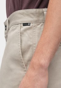 Μπεζ βαμβακερό παντελόνι με ίσια γραμμή, που διαθέτει μαύρη ετικέτα λογότυπου και κλείσιμο με κουμπί. Το ύφασμα έχει λεπτή υφή.