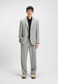 Helles graues Nadelstreifenanzug mit strukturiertem Blazer und weitem Hosenbein, kombiniert mit einem schwarzen Poloshirt und beigen Schuhen.
