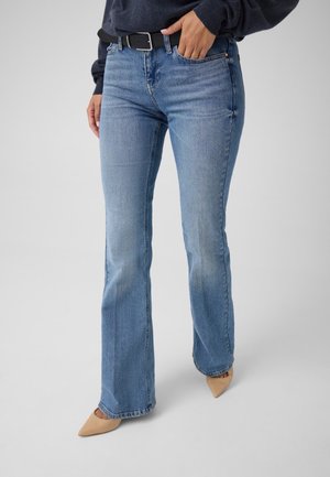 Personne portant un jean bootcut bleu clair, une ceinture noire, un haut foncé et des chaussures beiges à bout pointu, debout avec une main dans la poche.