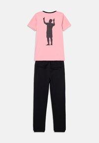 Maglietta rosa con grafica nera di una figura in piedi sul retro, abbinata a pantaloni felpati neri. Materiale in cotone, vestibilità comoda.