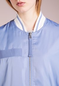 Primer plano de una persona que lleva una chaqueta de cremallera azul claro con un collar a rayas blancas y azules y una camiseta gris debajo.