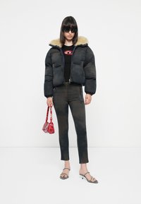 Zwarte pufferjack met een nepbontkraag, gecombineerd met donkere, slim-fit jeans en open teen sandalen. Accessoire: kleine rode handtas.