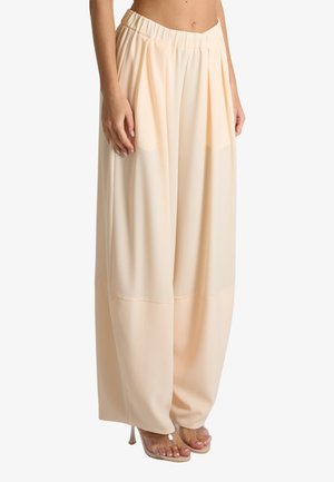 Donna che indossa pantaloni beige chiaro a vita alta, gamba larga, con pieghe e fascia elastica in vita, abbinati a décolleté trasparenti e aperte sulla punta.