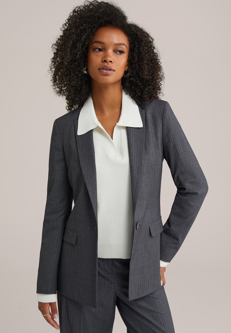 Šedý pruhovaný blazer s klopovým límcem, zkombinovaný se světle šedým žebrovaným svetr. Materiál se zdá být hladký s pevnou krejčovskou úpravou.