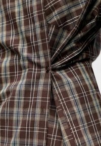 Chemise à carreaux brune présentant un mélange de lignes beige et marine, avec un devant boutonné et une poche unique en tissu texturé.