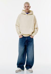 Beige oversized huva-tröja, vida blå jeans och vita sneakers. Slät tyg, avslappnad passform, med en detalj för framfickan.