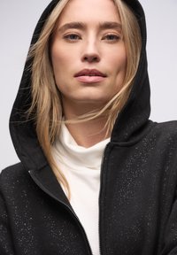 Czarna bluza z kapturem na suwak, z fakturowanym wzorem z rozproszonymi białymi plamkami. Noszona na białym golfie, z widocznym kapturem.