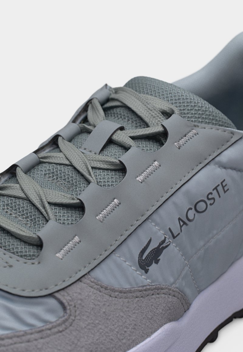 Nahaufnahme eines grauen Lacoste-Sneakers mit strukturiertem Stoff, synthetischen Überzügen, Schnürdesign und dem Markennamen mit Krokodil-Logo an der Seite.