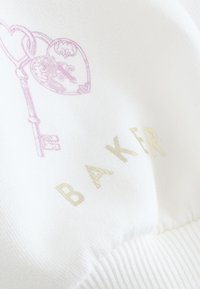 Illustration délicate d’un cadenas et d’une clé roses en forme de cœur au-dessus du texte doré « BAKER » sur un tissu blanc doux avec bord côtelé.