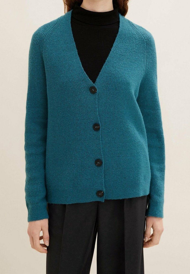 Femme portant un cardigan boutonné bleu sarcelle sur un col roulé noir et un pantalon noir, debout devant un fond uni.