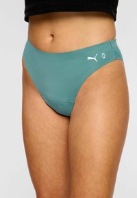 Femme portant des bas de bikini teal à taille haute de Puma et un haut court noir, montrant le milieu du ventre et le haut des cuisses.