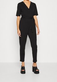 Mono negro con diseño envolvente, mangas cortas, cinturilla ajustable y piernas acampanadas. Combinado con sandalias de plataforma negras y gruesas.