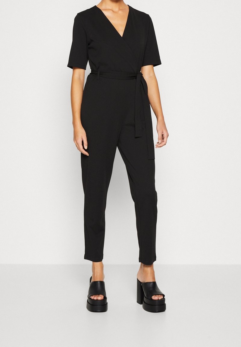 Mono negro con diseño envolvente, mangas cortas, cinturilla ajustable y piernas acampanadas. Combinado con sandalias de plataforma negras y gruesas.