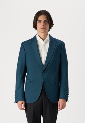 Türkisfarbener Blazer mit strukturiertem Finish, Reverskragen, zwei Knöpfen und Taschen. Kombiniert mit einem weißen Hemd und dunklen Hosen.