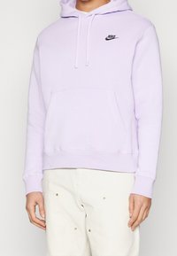 Person som bär en ljuslila Nike-hoodie med ficka framtill och vita byxor, visad från axlarna till höfterna mot en enfärgad bakgrund.