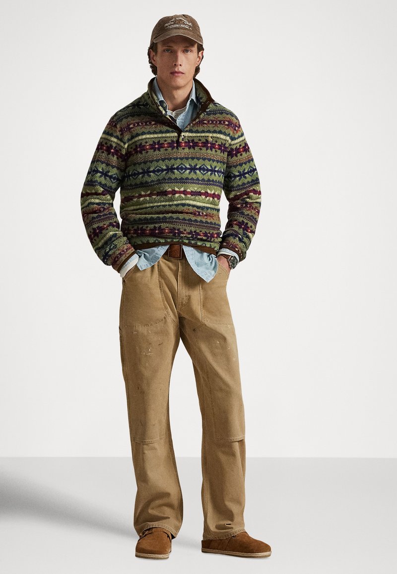 Pull tricoté texturé multicolore avec col montant sur une chemise bleu clair, assorti à un pantalon beige et des chaussures plates marron.