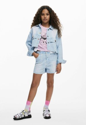 Chaqueta y pantalones cortos de mezclilla azul claro con bordes deshilachados, combinados con una camiseta gráfica rosa y sandalias plateadas con calcetas rosas y amarillas.