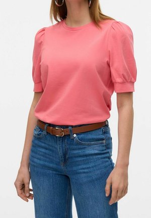 Vrouw die een koraalroze t-shirt met pofmouwen draagt, ingestopt in een blauwe jeans met een bruine riem, staand met ontspannen handen langs haar zij.