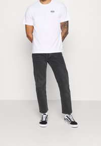 Bílá bavlněná tričko s malým modrým logem, kombinované s tmavě šedými džíny a černobílými vzorovanými teniskami. Hladký materiál, volný střih.