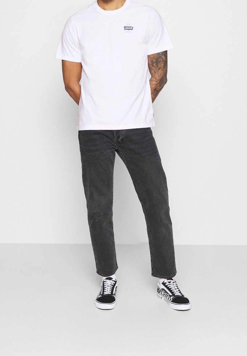Bílá bavlněná tričko s malým modrým logem, kombinované s tmavě šedými džíny a černobílými vzorovanými teniskami. Hladký materiál, volný střih.