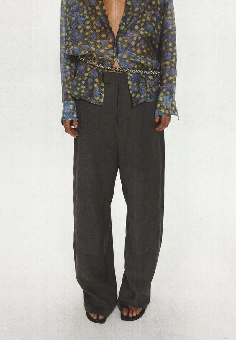 Personne portant un pantalon ample et sombre, un chemisier fleuri ouvert avec un motif jaune et bleu, une ceinture chaîne et des sandales noires à bouts ouverts.