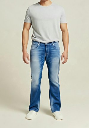 Male wearing light gray short-sleeve t-shirt, blue faded jeans, and white shoes, standing against a plain beige background.