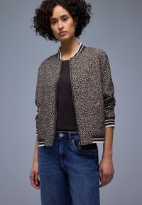 Donna con capelli ricci che indossa una giacca bomber a stampa leopardata sopra una maglietta nera e jeans blu, in piedi davanti a uno sfondo semplice.