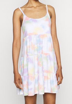 Kobieta w bezrękawowej, wielowarstwowej mini sukience z pastelowym wzorem tie-dye i cienkimi ramiączkami, stojąca na jednolitym tle.