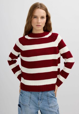 Joven mujer con cabello largo que lleva un suéter de punto a rayas rojas y blancas y jeans azules, de pie con las manos en las caderas frente a un fondo simple.