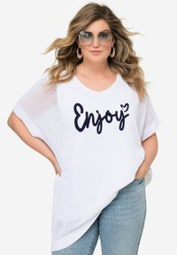 Top blanco de punto con diseño holgado, que presenta la palabra "Enjoy" en color navy. Combinado con jeans de mezclilla azul claro. Gafas de sol oversized transparentes.