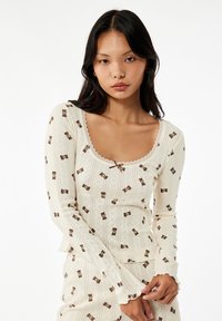 Cremefarvet langærmet top med teddy-bear mønster, bølget udskæring og struktureret stof. Har tætsiddende design og flagrende ærmer.