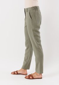 Pantaloni su misura verde oliva chiaro con pieghe frontali, cintura elastica e tasche laterali. Indossati con sandali piatti marroni. Tessuto dalla superficie liscia.