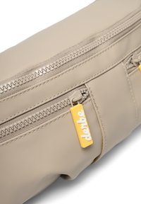 Beige taske lavet af glat materiale, med to lynlåslommer, gule hardware-accenter og et mærketag med hvid skrift.