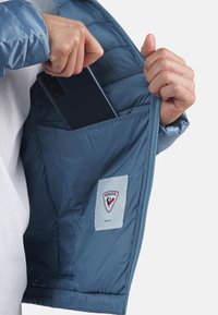 Vadderad blå jacka med en innerficka där en telefon delvis är insatt, som visar varumärkeslogotypen på en tyglapp.