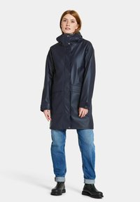 Marinblå vattentät regnkappa med hög krage, framknäppning och stora fickor, kombinerad med blå jeans och svarta ankelskor.