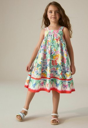 Noor tüdruk seisab, kandes värvilist suvekleiti troopiliste ja liblikate mustritega ning valgeid sandaale värviliste rihmadega.