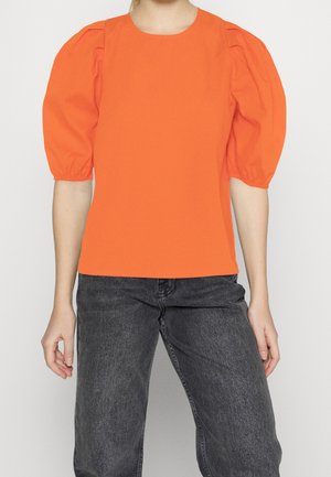 Kobieta ubrana w jasnopomarańczową bluzkę z bufkami i ciemnoszare dżinsy, stojąca na tle jednolitego jasnego tła.