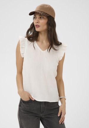 Femme portant une casquette beige avec un imprimé léopard sous la visière, un haut blanc sans manches à volants, un jean foncé et une montre, regardant vers la gauche avec la main dans la poche.