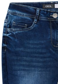 Jean en denim bleu foncé avec une texture lisse, poche avant, bouton argenté et détails de coutures subtiles. Étiquette de la marque visible sur la ceinture.