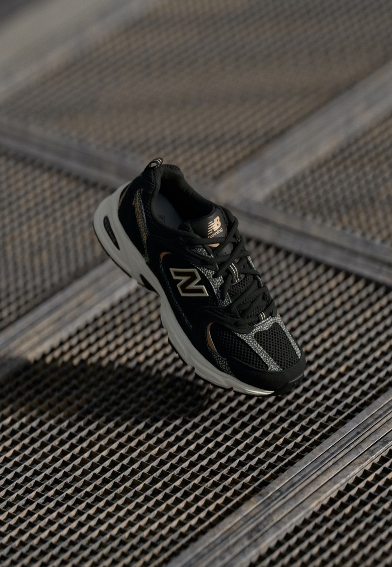Chaussure de course New Balance noire et grise flottant au-dessus d'une surface métallique texturée avec des motifs en forme de losanges.