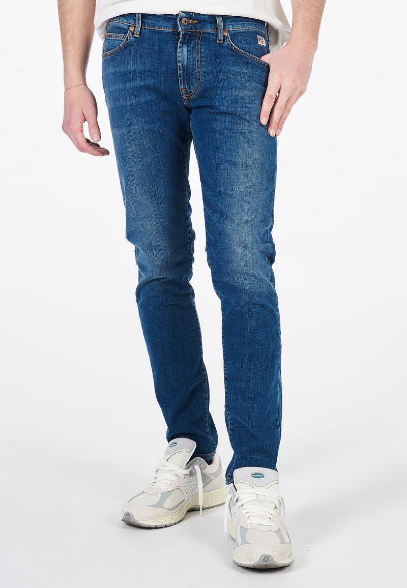 Jeans in denim blu con una vestibilità slim, caratterizzati da un leggero sbiadimento e dettagli di cucitura. Abbinati a scarpe sportive grigie e bianche.