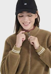 Braune Fleecejacke mit hohem Kragen und Reißverschluss, mit gestreiften Bündchen. Eine schwarze Mütze mit Logo wird getragen. Glatte Textur sichtbar.